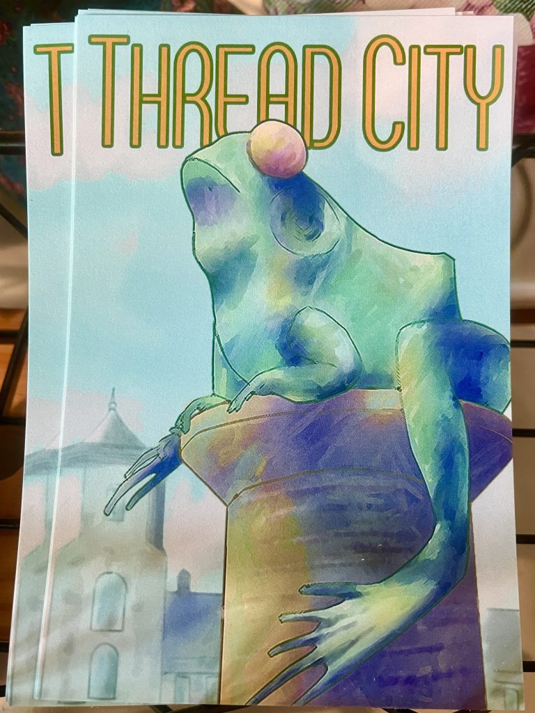 A stack of post cards displaying artwork depicting one of the sculptures of frogs sitting atop spools on Willimantic's "Frog Bridge"