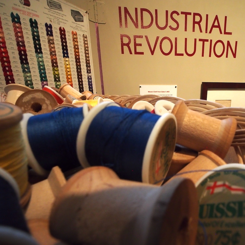 A close-up view of spools of sewing thread against a backdrop of a thread color chart and a sign that says "Industrial Revolution"